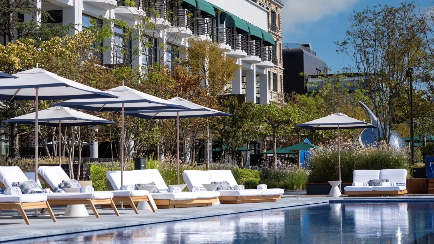 White loungers under parasols at the pool’s edge, with the gardens and the hotel’s façade and green awnings behind.