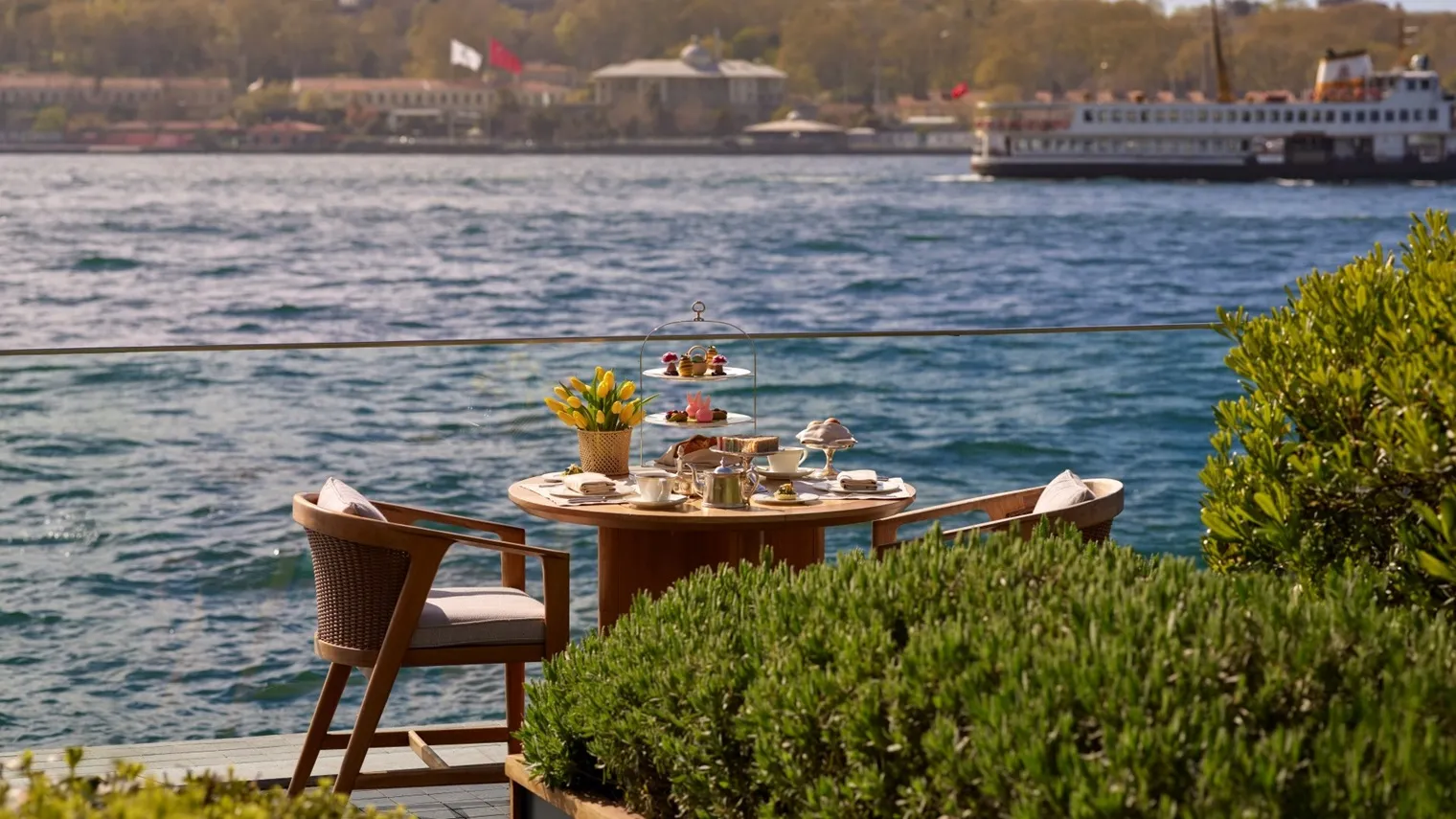 A table for 2 laid for afternoon tea in the garden, with a tiered stand and a vase of yellow tulips, on the edge of the Bosphorus; a ferry passes behind.