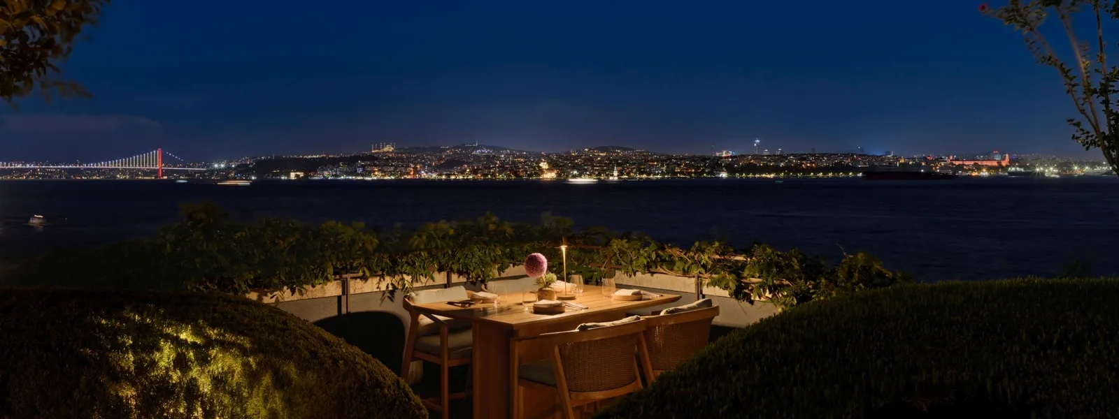 GALLADA’s terrace at night: a laid table among the hedges, the dark Bosphorus and, on the far shore, the city lights and a lit bridge.