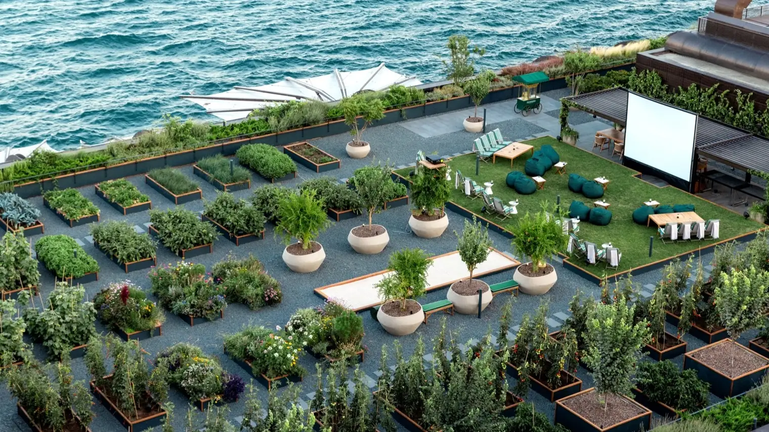 The rooftop herb garden from above: planted beds and potted trees, a lawn with beanbags and an outdoor cinema screen, and the Bosphorus just beyond.