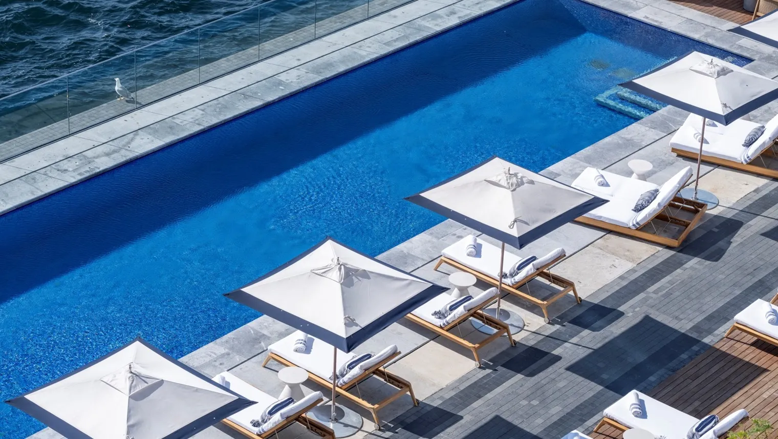 The outdoor pool from above: blue water along the glass rail over the Bosphorus and a row of loungers and white parasols on the deck.
