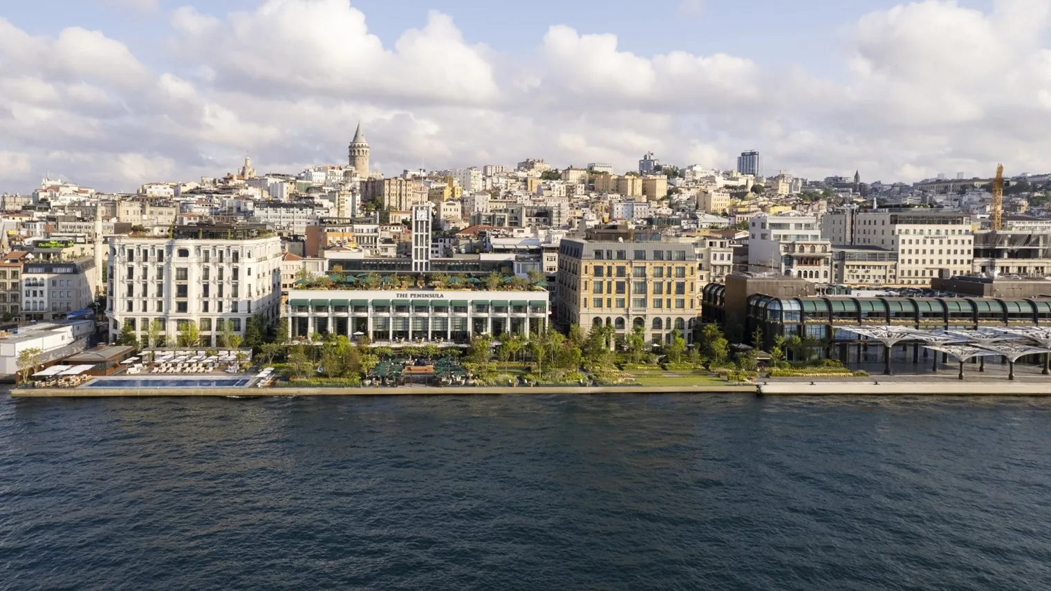 The hotel’s front by day, from the water: its buildings in a row on the Karaköy shore and the city climbing to the Galata Tower under a clouded sky.
