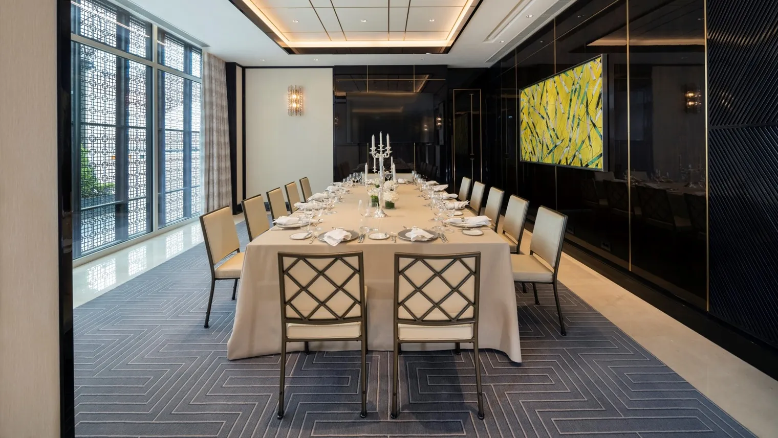 A meeting room laid for dinner: a long table under a pale cloth, lattice-backed chairs, a glossy black wall and windows behind fretwork screens.