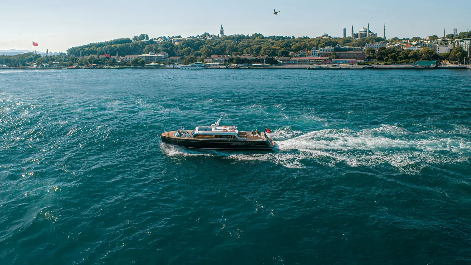 The yacht PEN 1 under way on the Bosphorus, seen from above, with the wooded headland of the old city, its domes and minarets, behind.