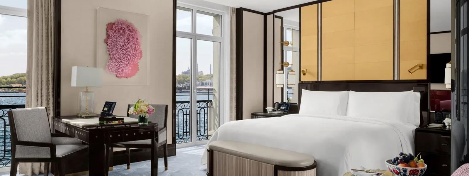 A Deluxe Bosphorus Room: the king bed with a bench at its foot, a desk with 2 chairs, a pink artwork on the wall and 2 French windows onto the water.