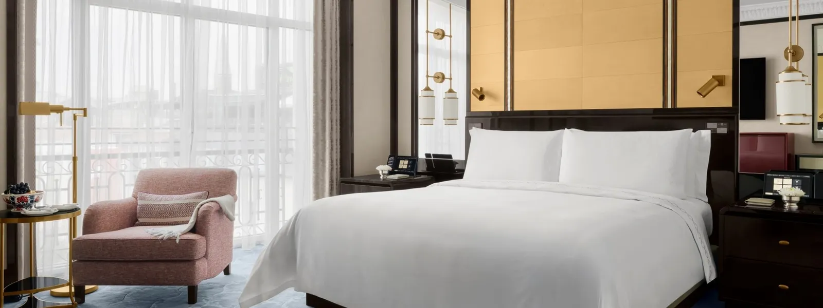 A Deluxe Room: the king bed, a pink armchair with a throw beside a reading lamp and the tall window veiled by sheers.
