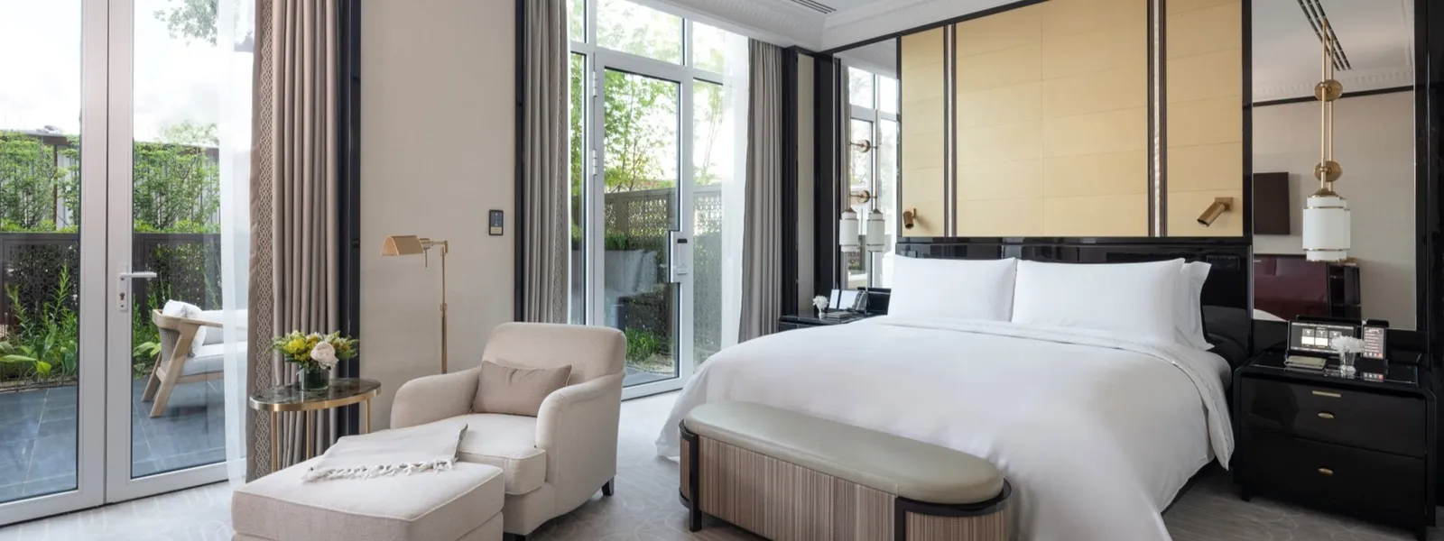 A Deluxe Suite with Garden bedroom: the king bed with its bench, an armchair and footstool and French windows opening onto the planted terrace.