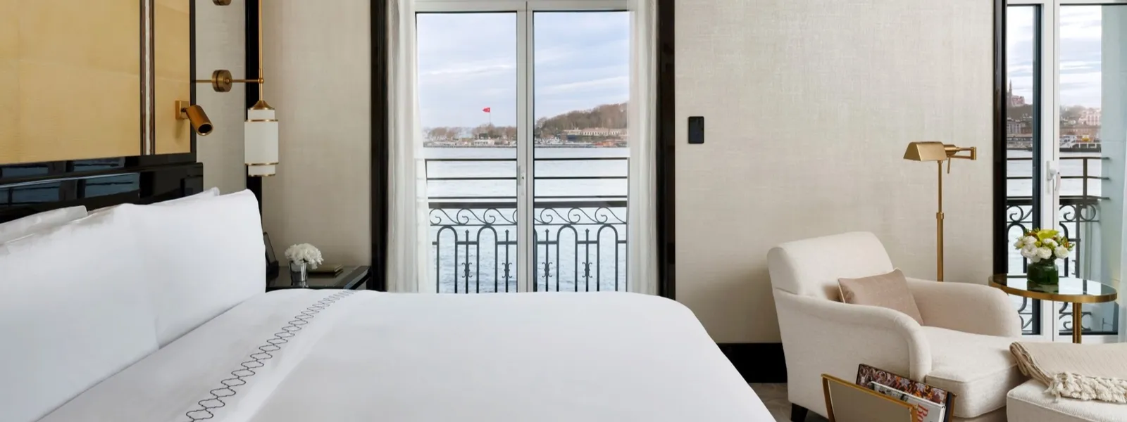 An Executive Bosphorus Suite bedroom: the bed in the foreground, a pale armchair and French windows with iron rails over the Bosphorus.