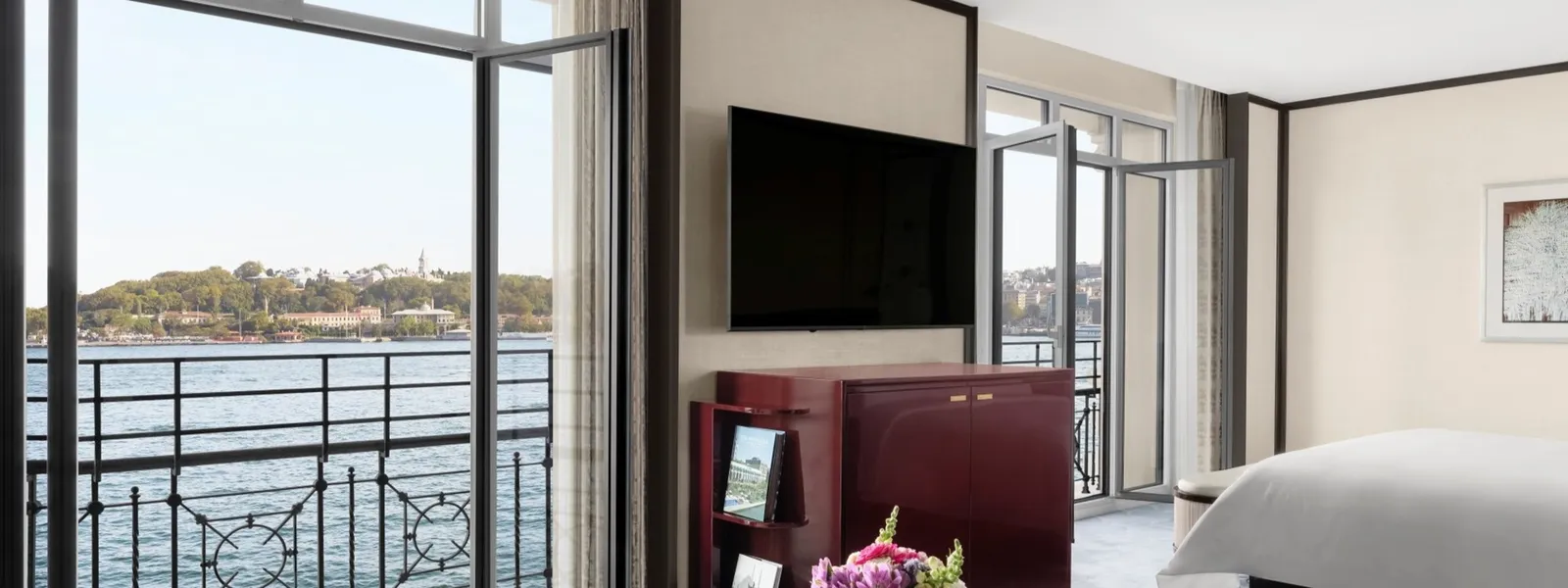 A Grand Deluxe Bosphorus Room: French windows open on the Bosphorus and the old city’s headland, the red lacquered cabinet under the television and a corner of the bed.
