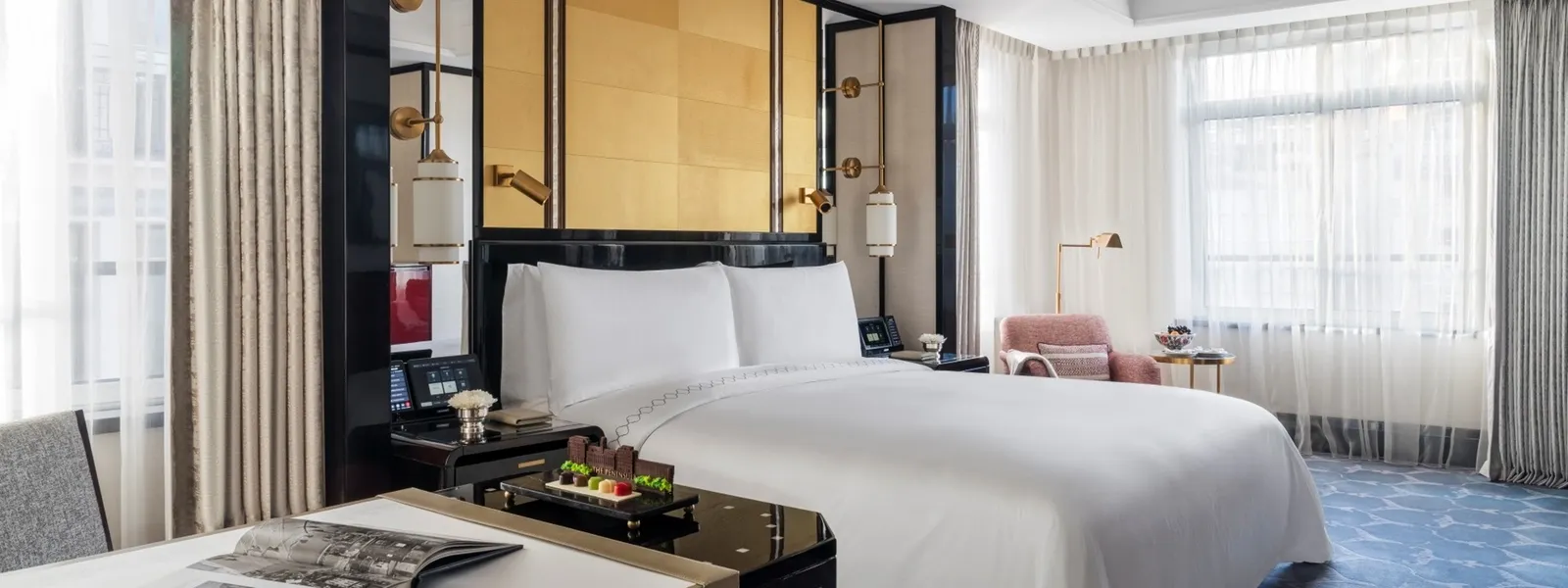 A Grand Deluxe Room: the king bed, windows on 2 sides, a pink armchair and, in the foreground, the desk with a tray and an open magazine.