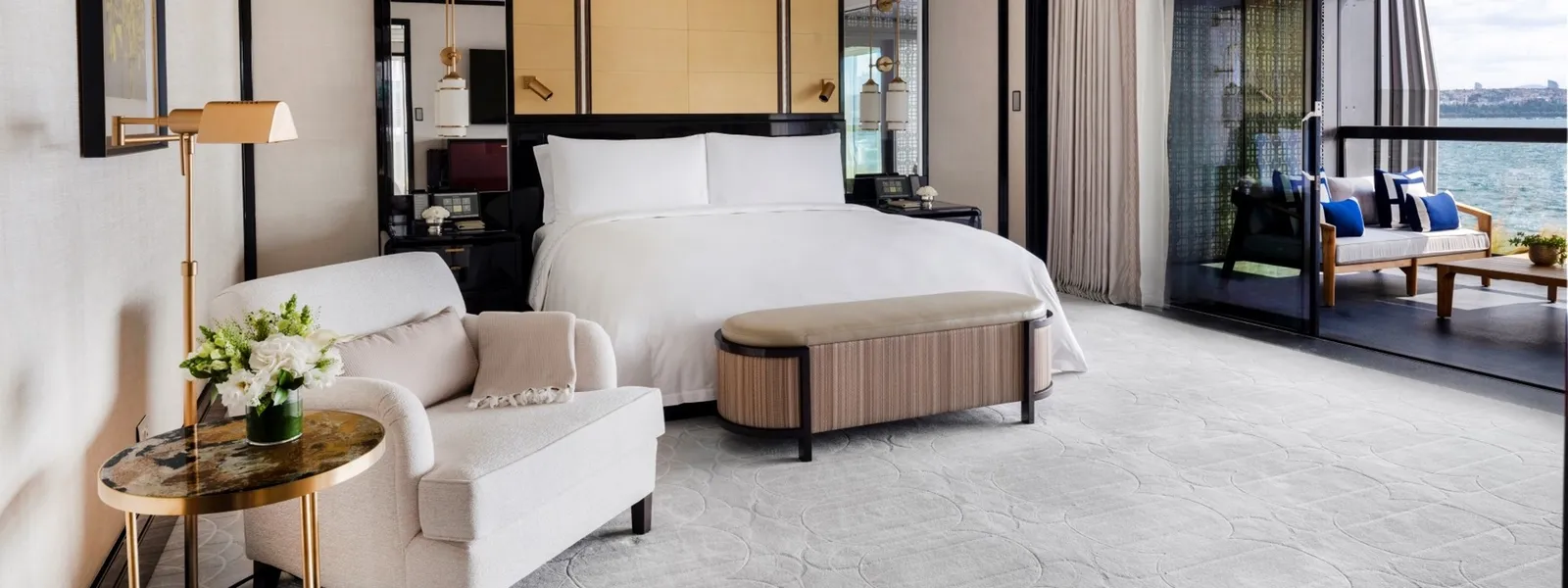 A Premier Bosphorus Suite with Balcony bedroom: the king bed with its bench, a pale chaise longue and the furnished balcony with blue cushions over the Bosphorus.