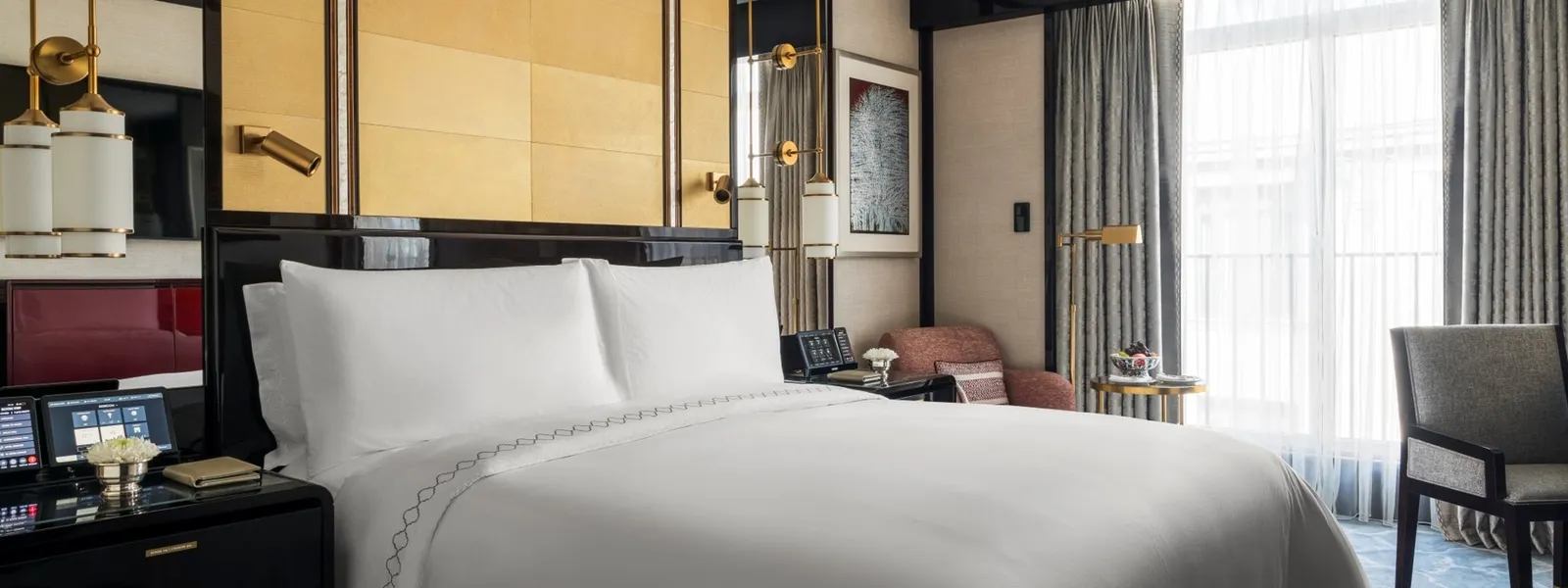 A Superior Room: the king bed against a gold-panelled headboard, dark lacquered bedside tables, a pink armchair and the window behind sheer curtains.