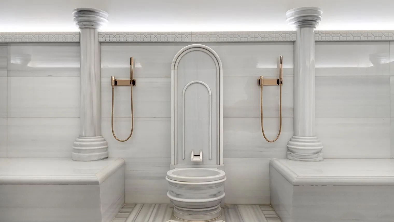 A hammam room: white marble walls and benches, 2 columns, a marble basin under an arched niche and brass hand showers.