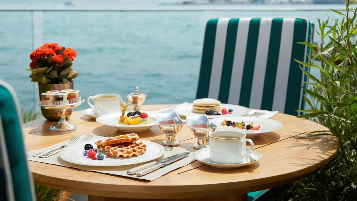 Breakfast on a round wooden table on the terrace: waffles with berries, pancakes, tea and jams, a green-and-white striped chair and the Bosphorus beyond the glass rail.