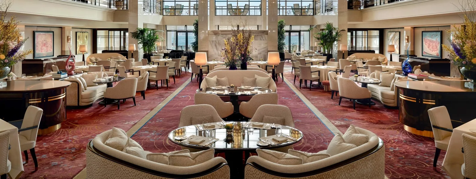 The Lobby: pale sofas and armchairs around tables on a red carpet, tall flower arrangements and double-height windows at the far end.