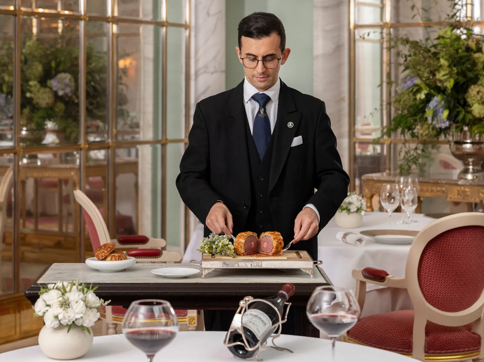 A waiter in a dark suit carves the Beef Wellington at the trolley beside a laid table
