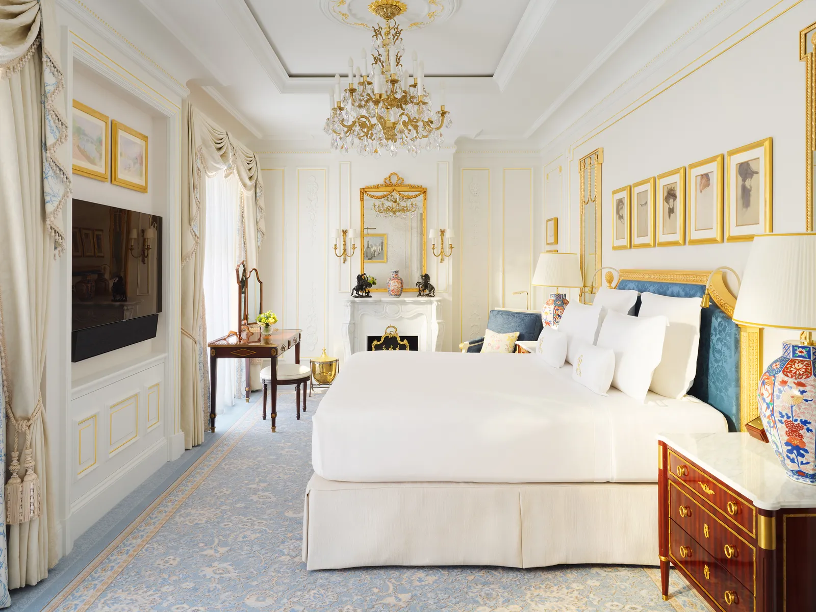 Una Deluxe Room: letto bianco, pareti avorio con quadri dalle cornici dorate, un vaso blu e bianco e un lampadario