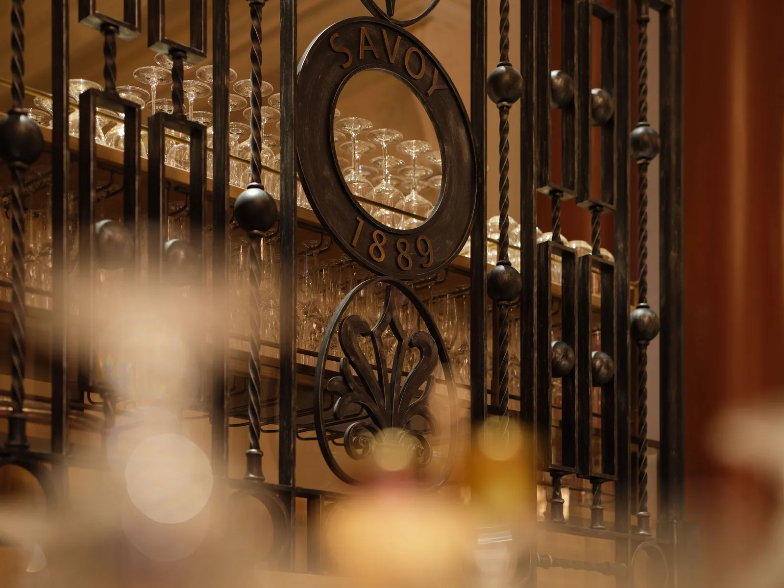 A wrought-iron grille with the “Savoy 1889” medallion before the lights of the bar in Gallery
