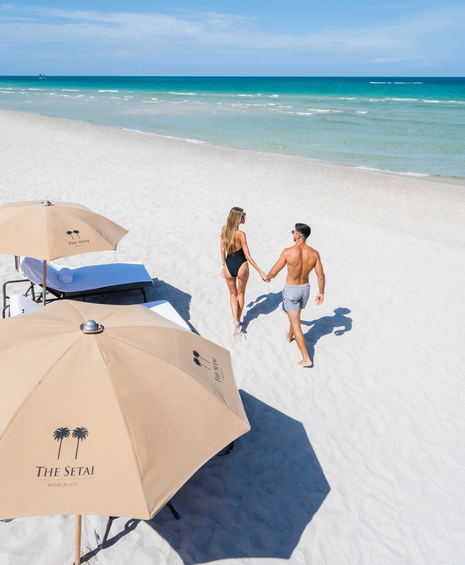 A couple walk hand in hand across the white sand towards the sea, past 2 umbrellas bearing the hotel’s logo.