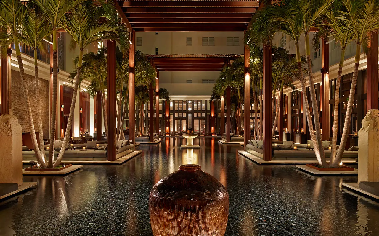 The courtyard at night: the dark reflecting pond with a large jar at its centre, lit palms and timber pergolas.