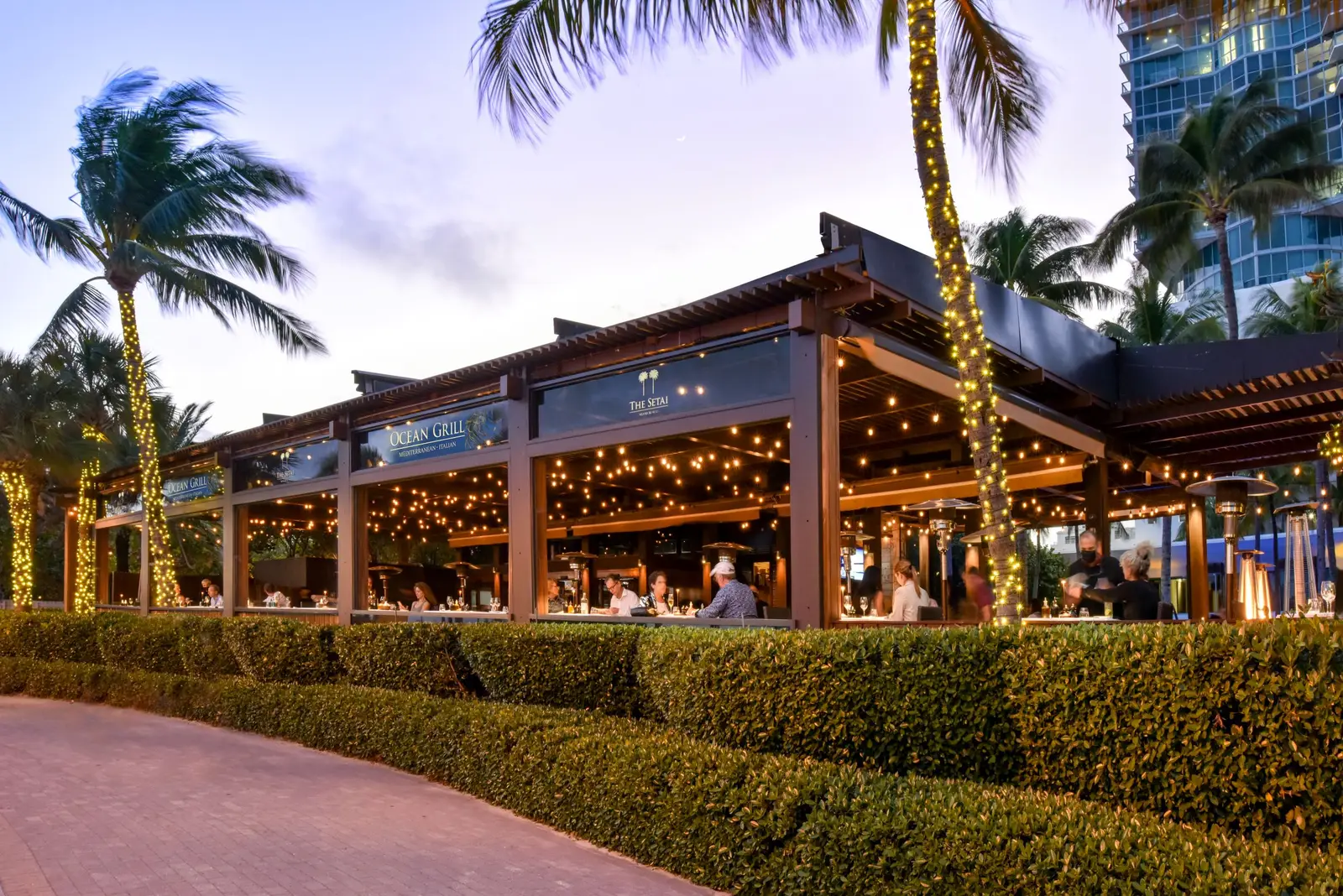 Ocean Grill at dusk, an open pavilion with its lights on behind a hedge, among palms bent by the wind.