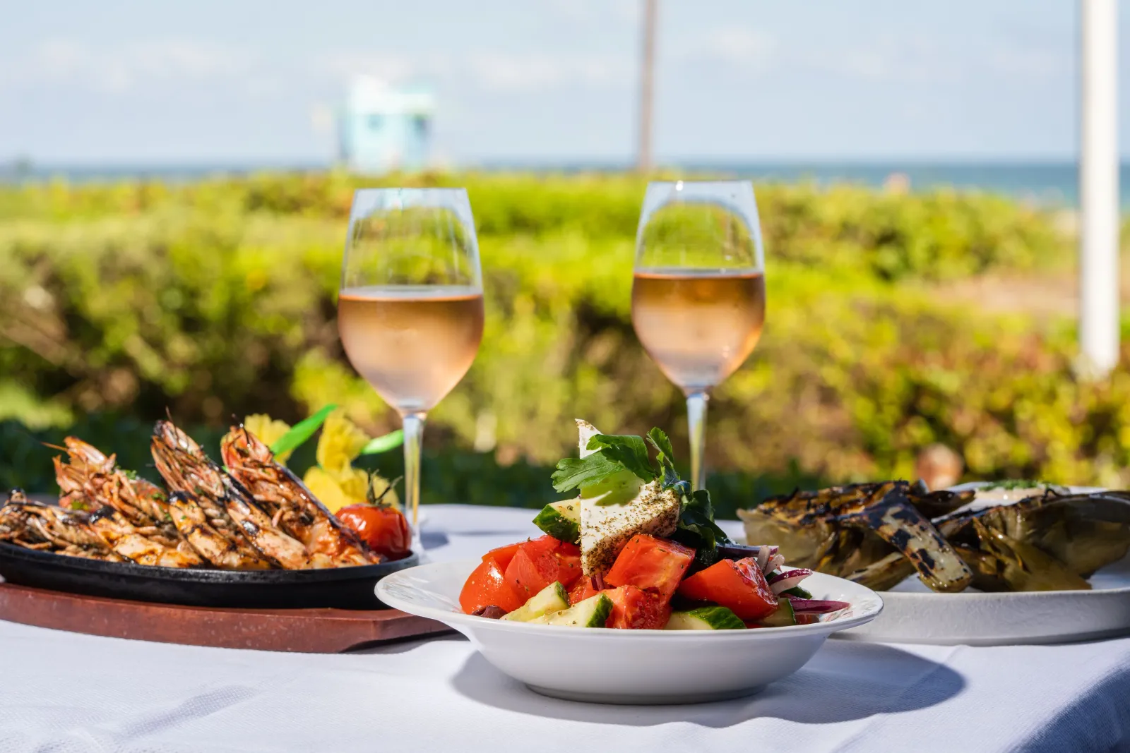 2 glasses of rosé, grilled prawns and a Greek salad on a table at Ocean Grill, the dunes and sea blurred behind.
