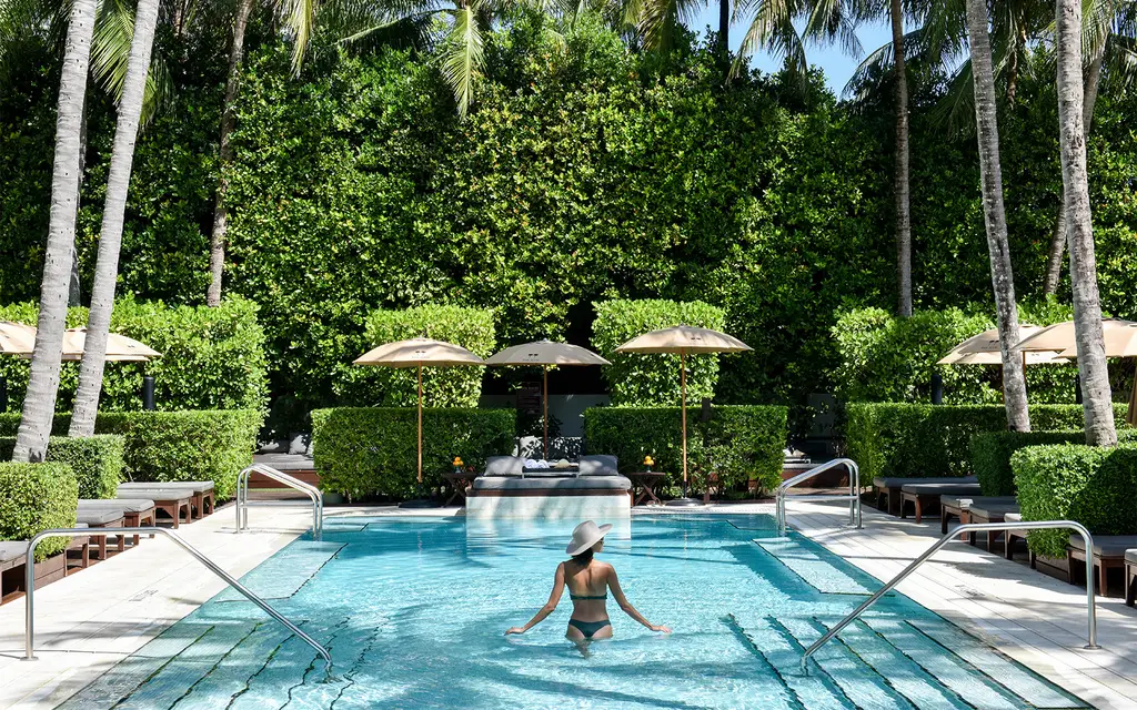 A woman in a hat wades into one of the pools from its steps, between tall hedges, palms and umbrellas.
