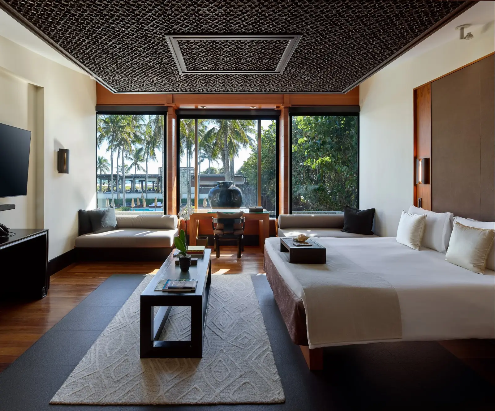 A Pool Villa: the low bed, a pale rug and 3 windows onto the palms and the pools, under a carved ceiling.