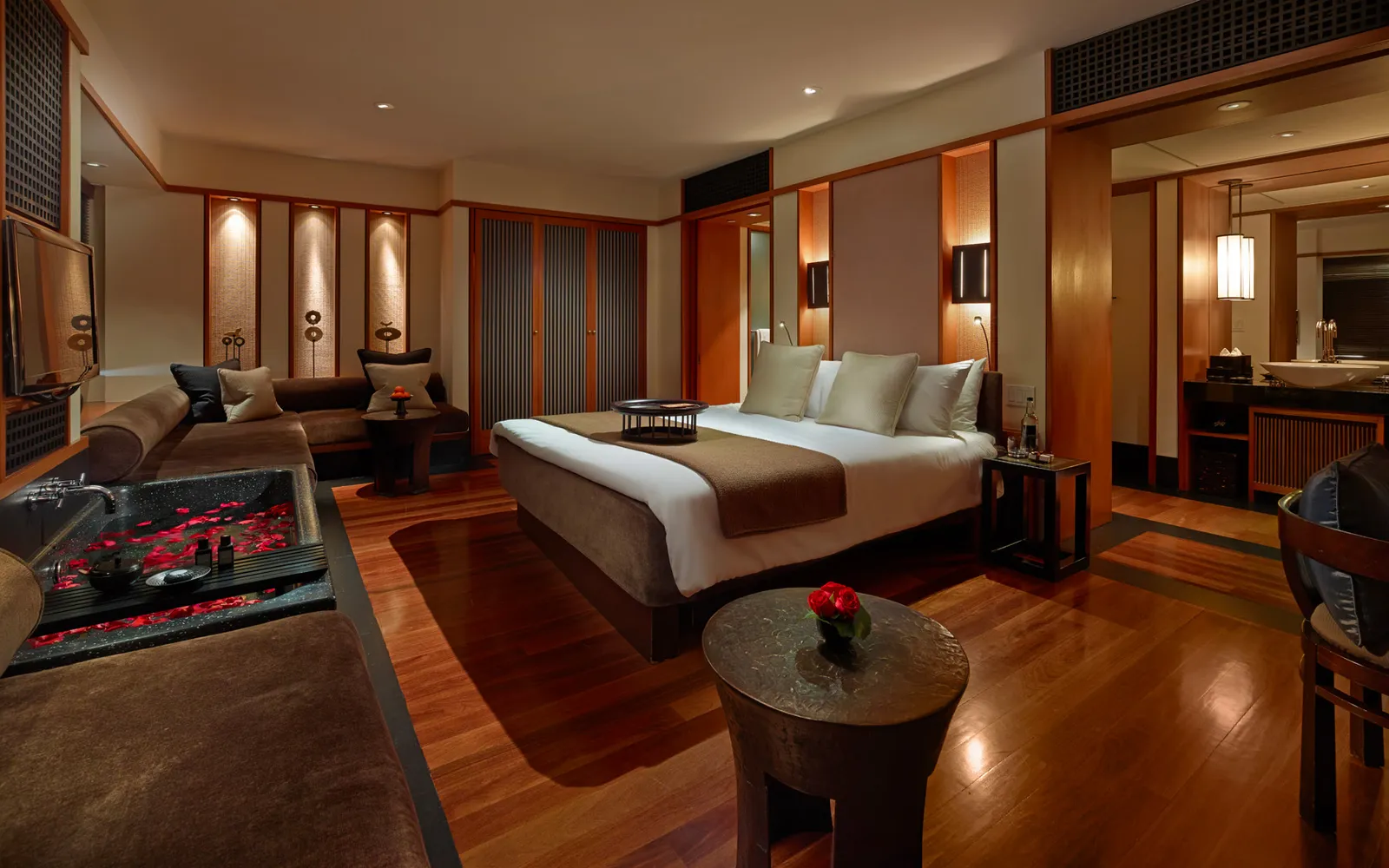 A Studio Suite in the evening: the bed, the black bath strewn with rose petals beside the sofa, and teak walls.