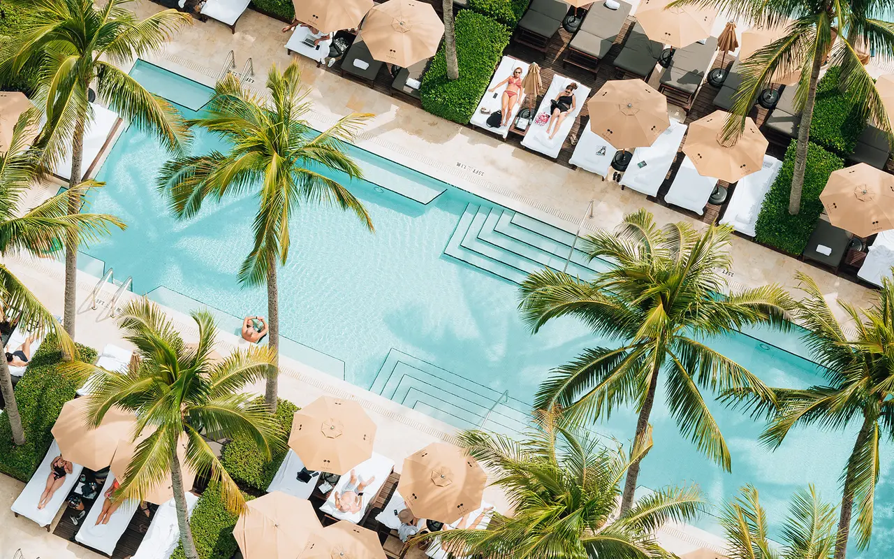 A pool seen from above, set diagonally among the palms, with pale umbrellas and guests on loungers.