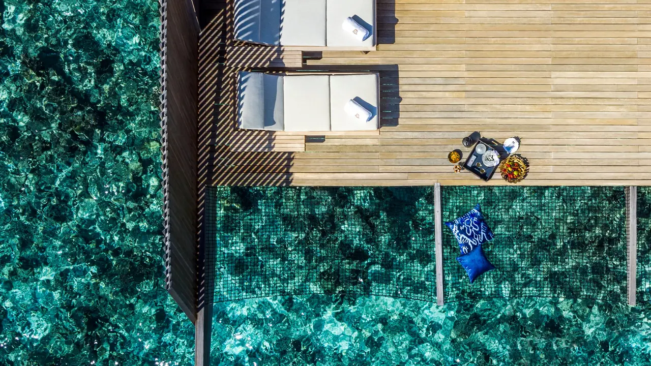 An overwater villa deck seen from above, its loungers over the turquoise lagoon