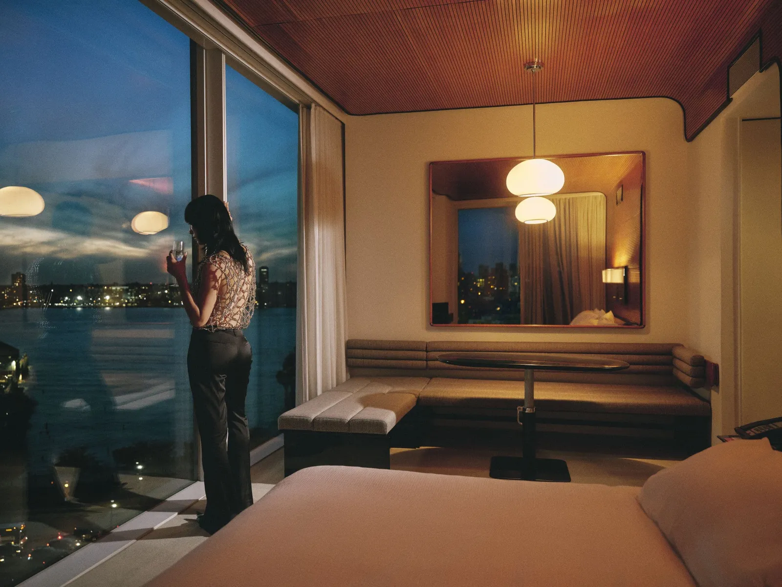 A room at night, a woman at the glass above the lights of the Hudson, the banquette and lamps lit