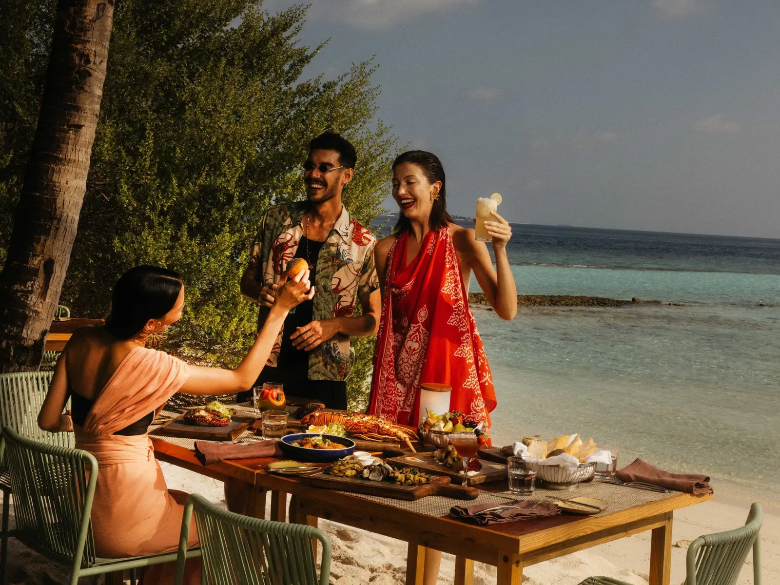 3 guests laughing at a table of grilled food on the BBQ Shak’s beach, the sea behind them.