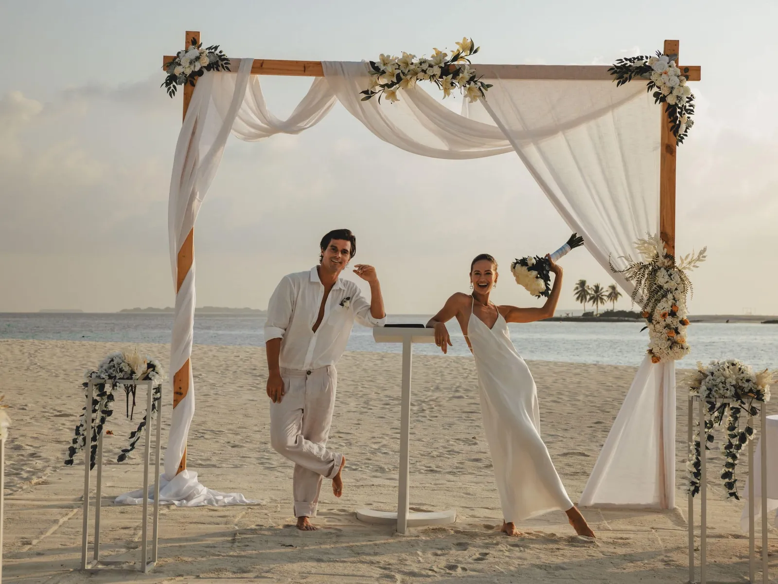 A couple dressed in white under an arch of flowers and drapes on the beach.