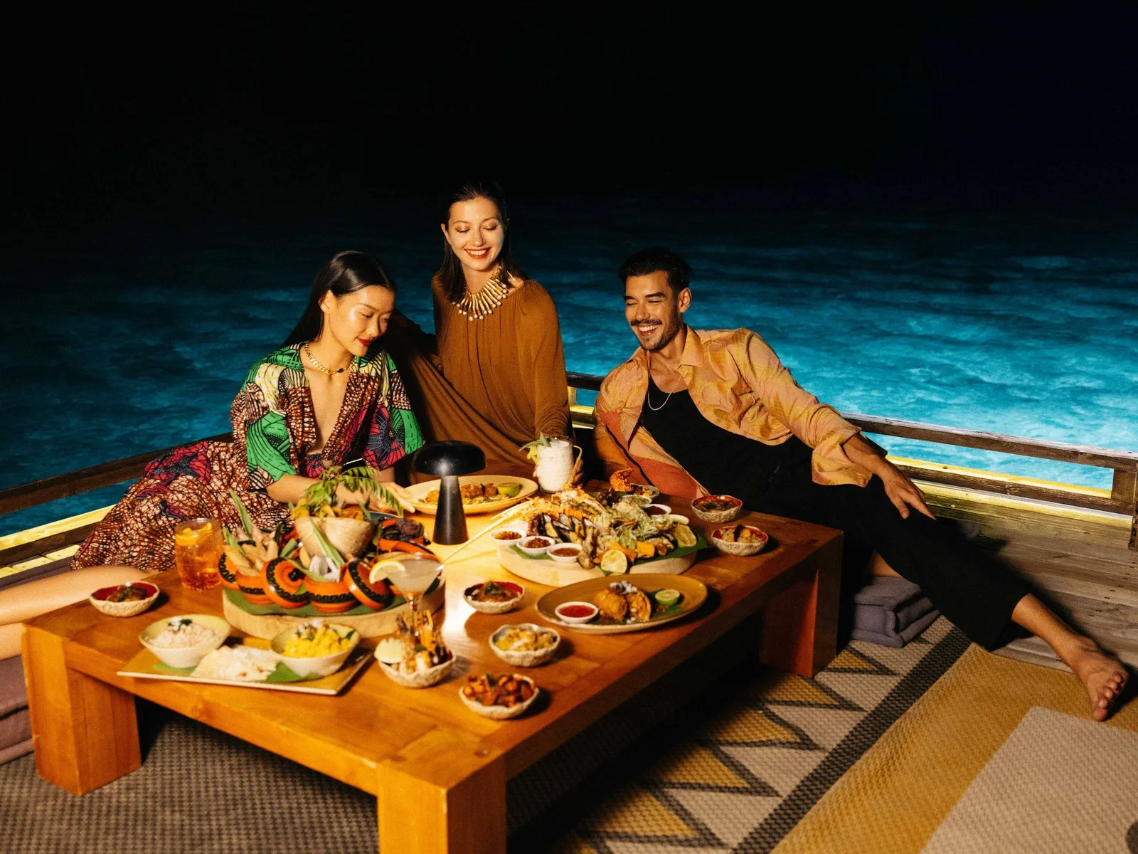 Dinner at Guduguda: 3 guests sitting low on cushions round a table full of dishes, the dark sea behind them.
