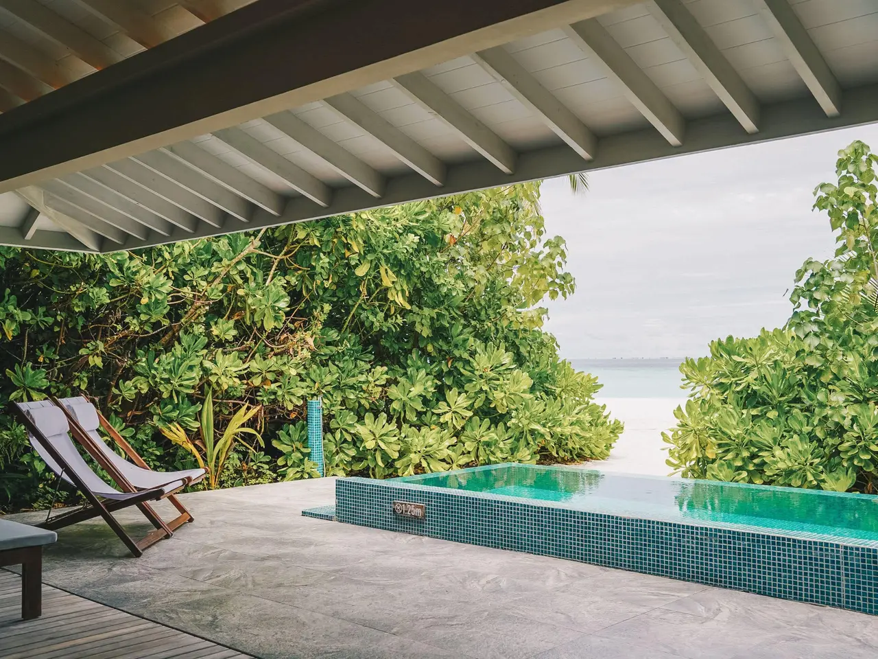 A beach villa’s covered deck, with its infinity pool, a deckchair and the sand between the greenery.