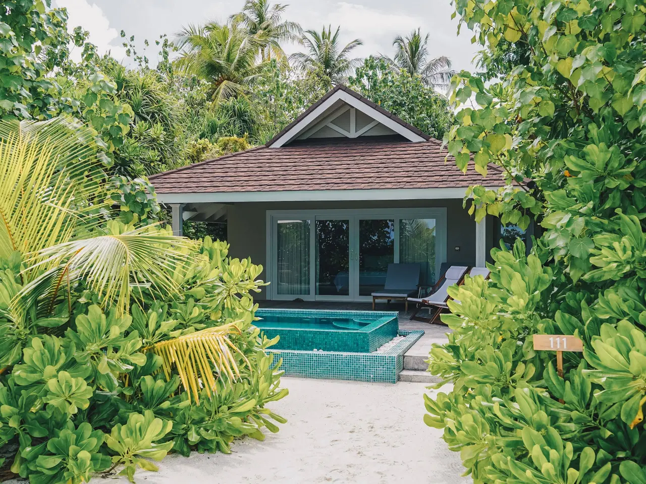 A beach villa among palms and greenery, its pool in front of the glass doors and the loungers.