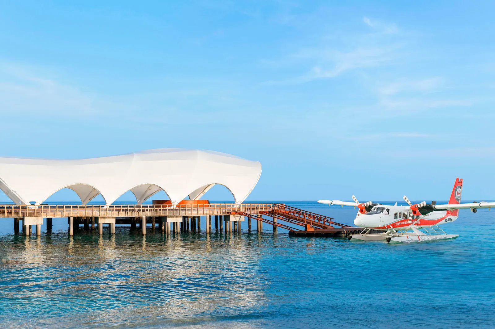 The seaplane moored at the arrival jetty, under the large white shell-shaped canopy, with a flat sea and a blue sky.