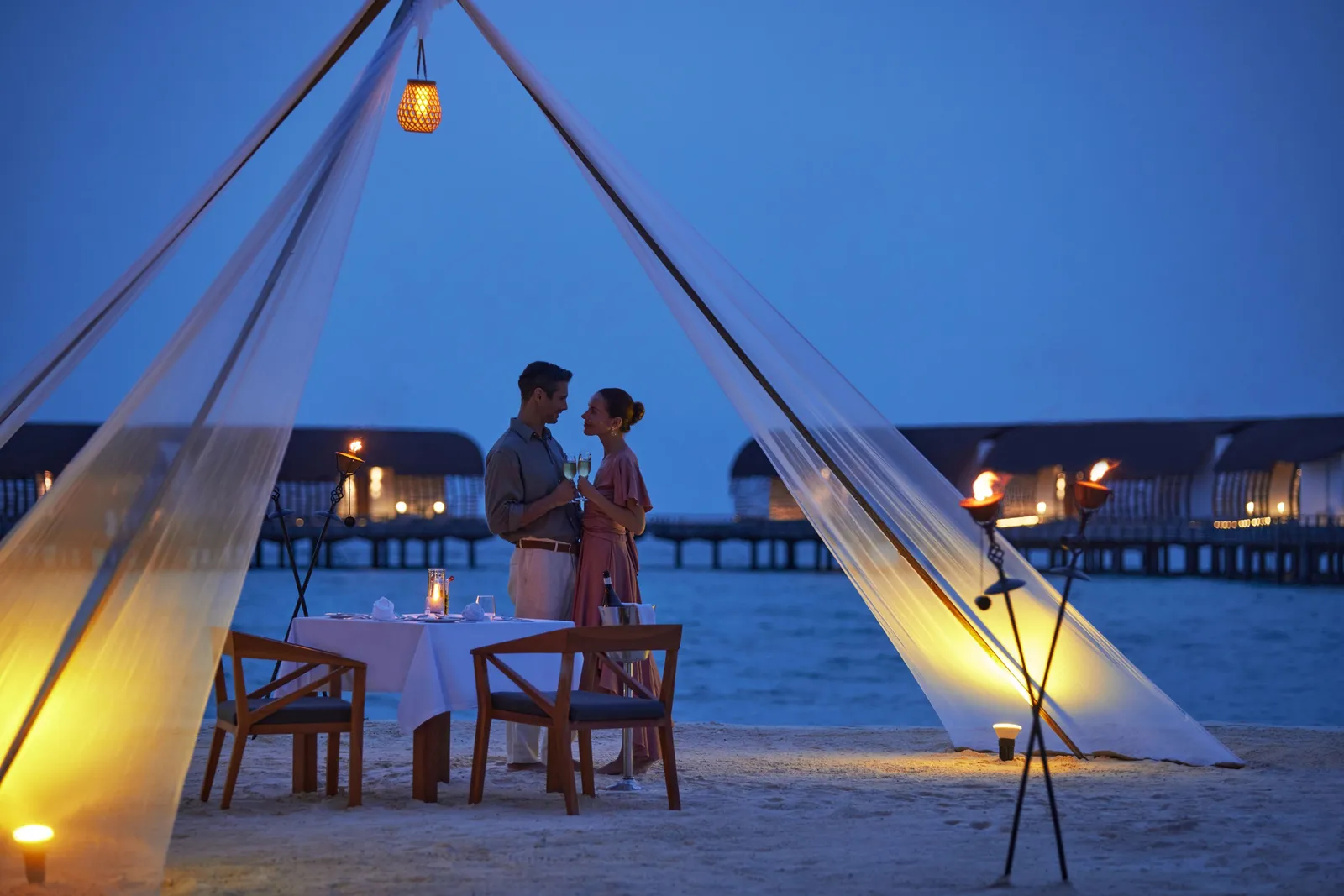 Dinner for 2 on the beach at night, under a pointed white canopy lit by torches, with the water villas behind.