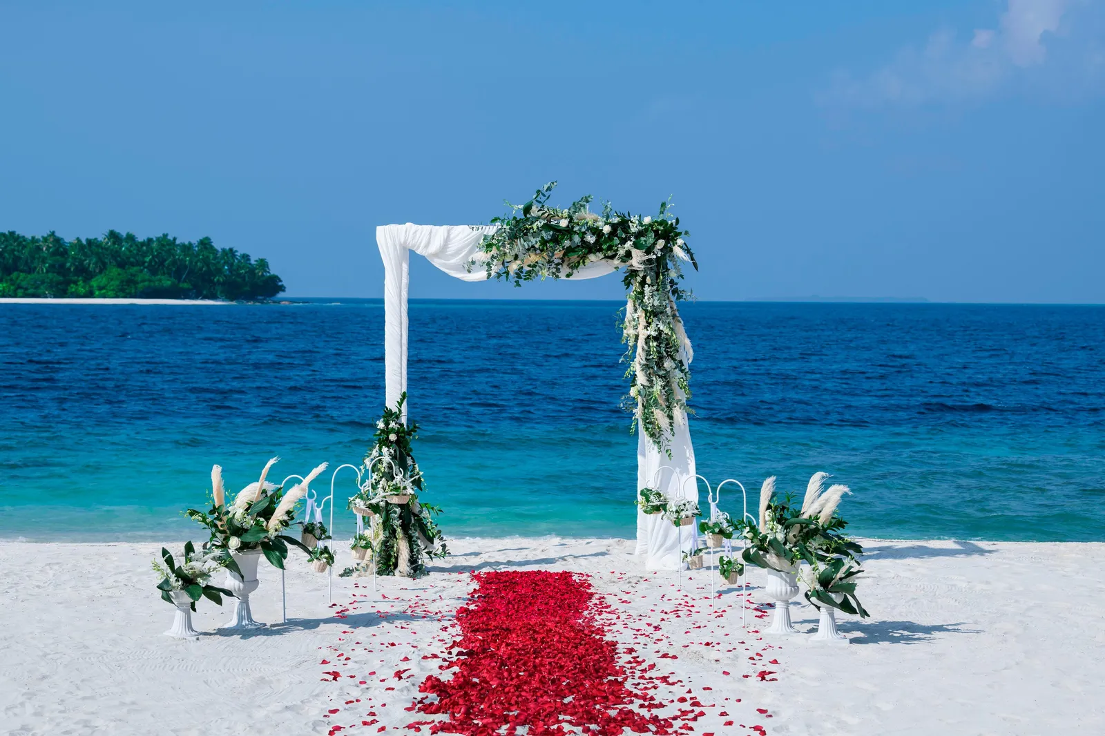 An arch of white flowers on the beach facing the sea, with a path of red petals on the sand.