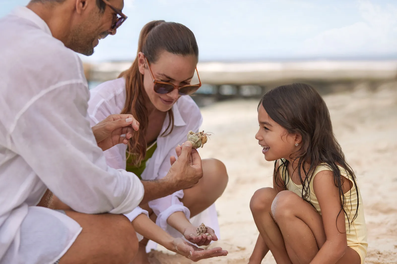 2 parents and a little girl crouched on the sand look at a small hermit crab in the mother’s hand.