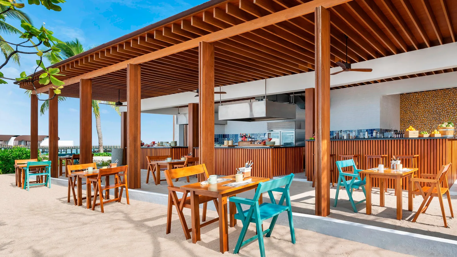 Hawker by day: tables and coloured chairs on the sand under the timber pergola, with the open kitchen at the back.