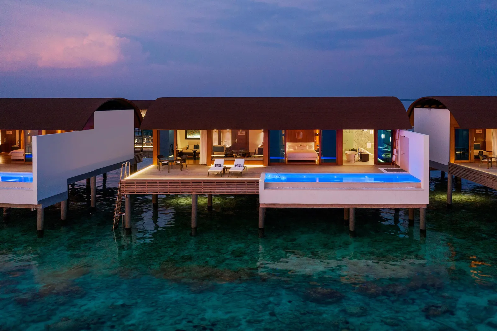 A water villa at dusk: the lit deck, the blue private pool and the ladder down into the lagoon.