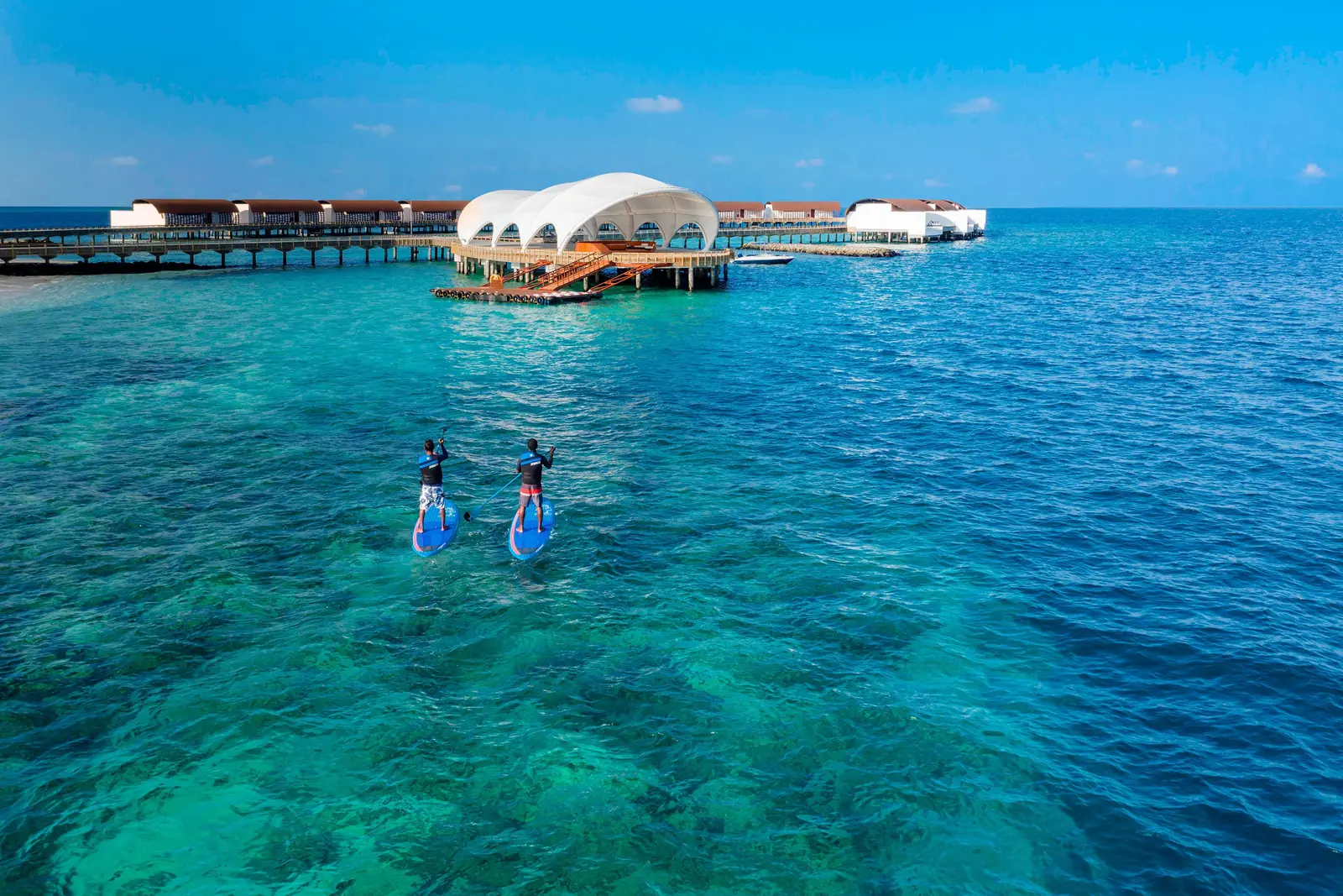 2 guests on paddleboards in clear water over the reef, with the arrival jetty and the water villas behind.
