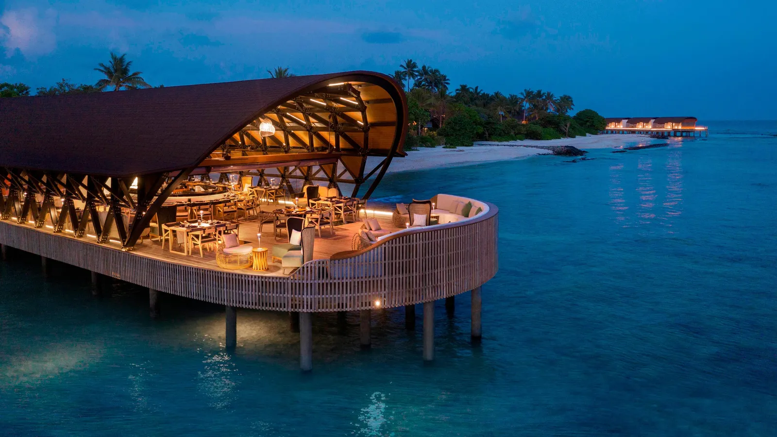 The Pearl in the evening, the dining room under its timber vault on stilts over the sea, lit up, with the beach behind.
