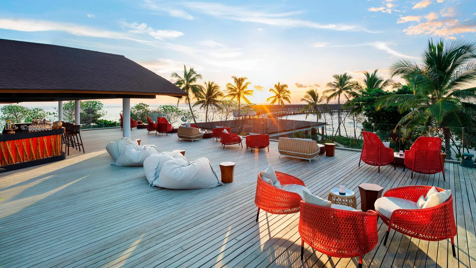 Sunset Bar’s terrace at sunset, with red armchairs, white beanbags and the sun dropping behind the palms.