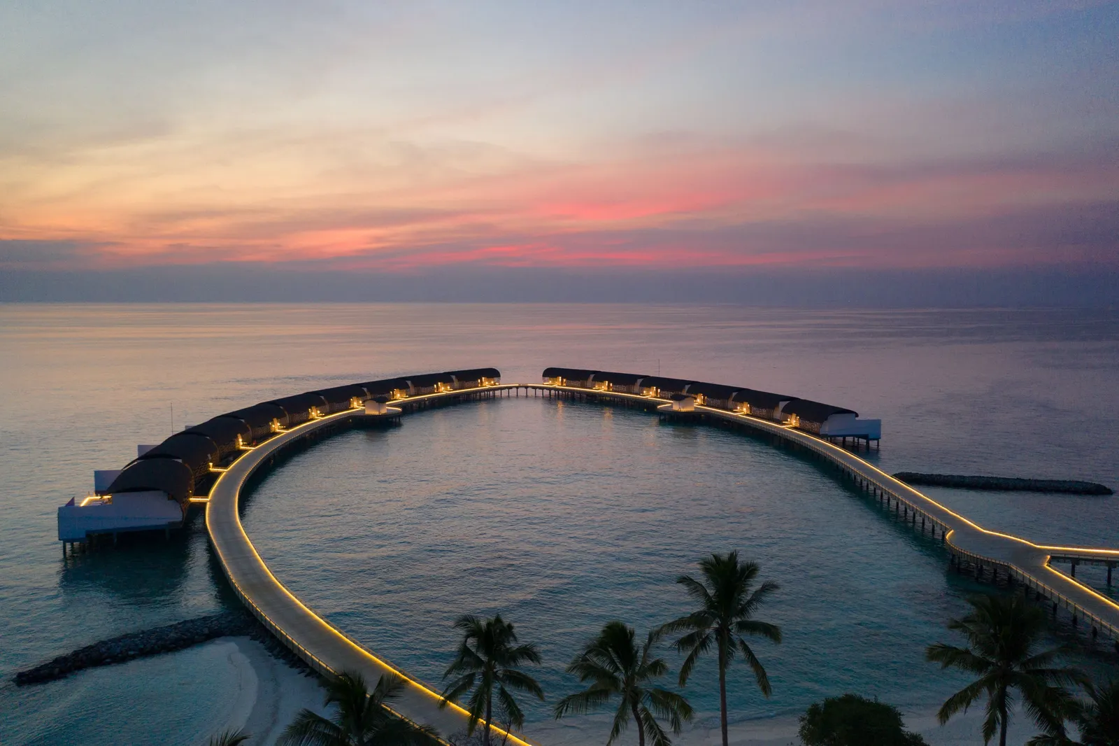 The jetty of water villas at sunset, a ring of lights over the sea with palms in the foreground.
