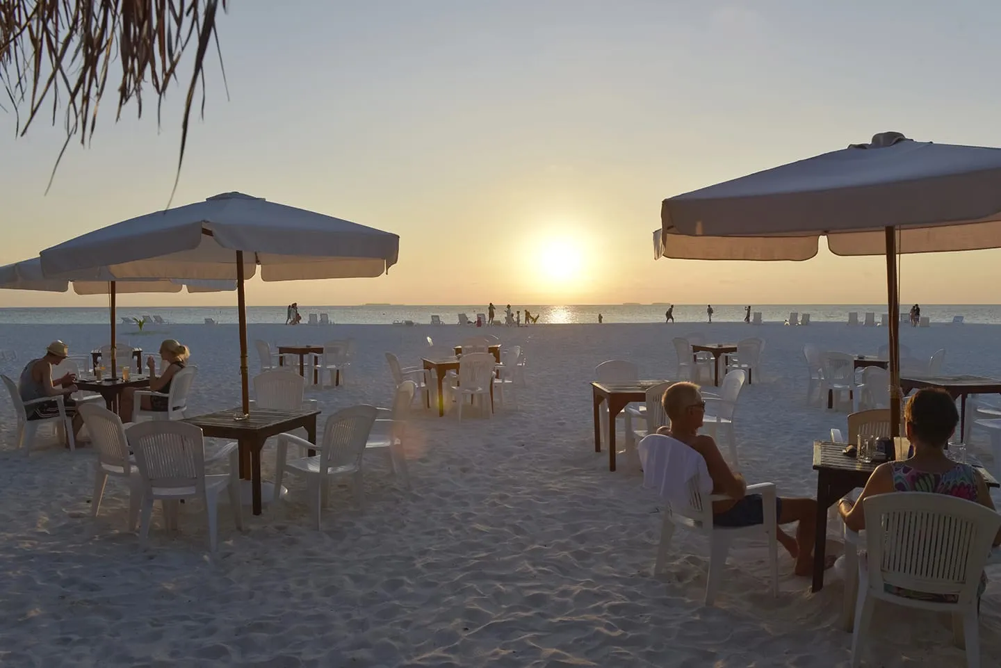 The beach bar’s tables at sunset under parasols, a few guests seated on the sand.
