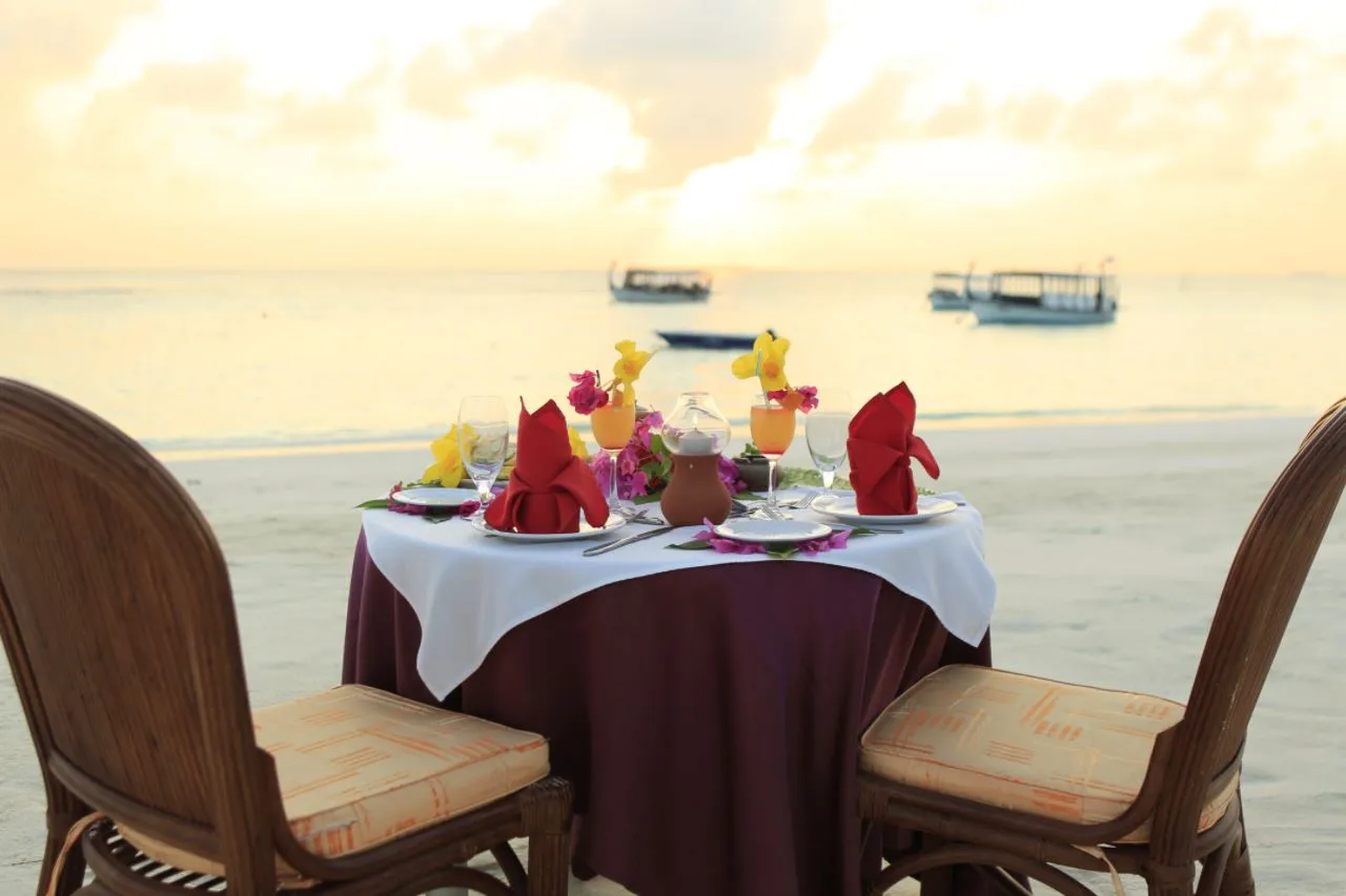 A table for 2 laid on the beach, with cocktails and flowers, and 2 dhonis on the sea behind.