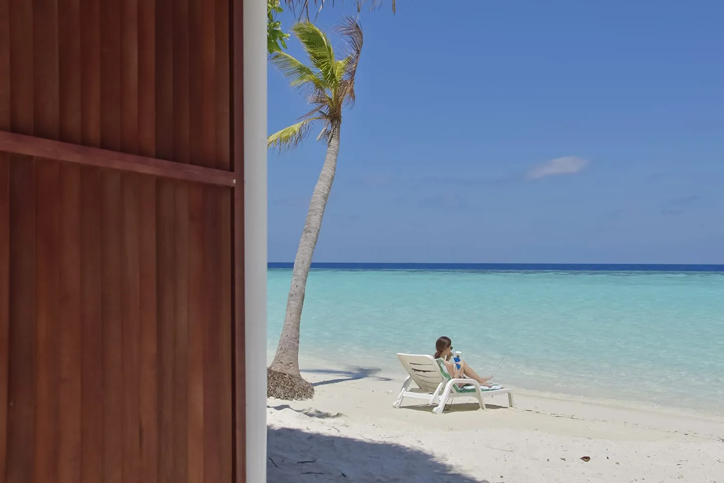 A woman on a lounger under a palm on the beach, seen from a bungalow’s wooden door.